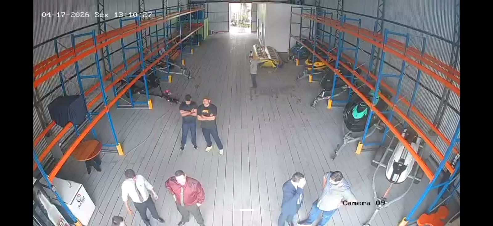 Cerca de três minutos depois. Galpão parcialmente esvaziado: prateleiras visivelmente sem embarcações, e o grupo ainda em deslocamento dentro do espaço.