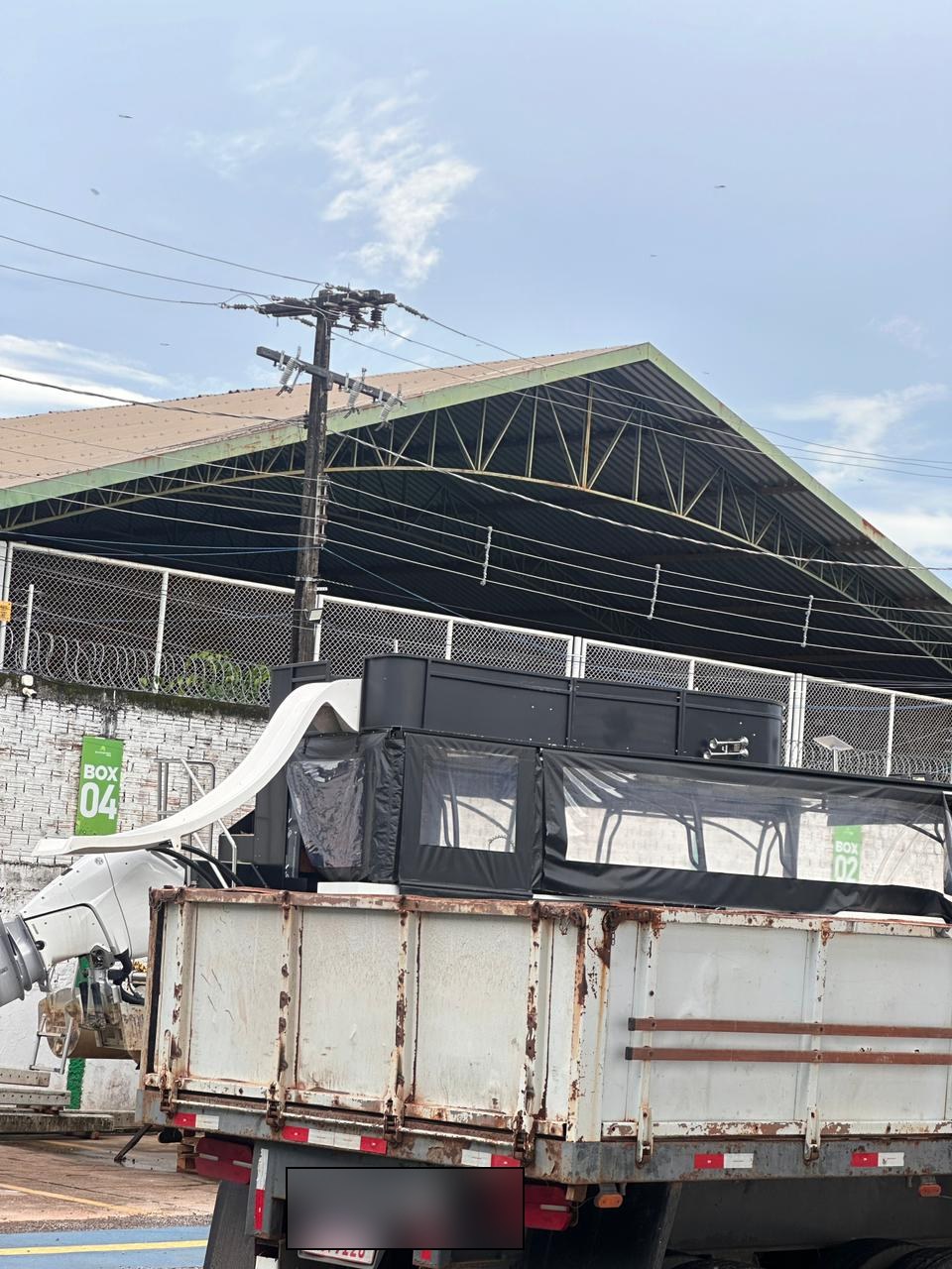 Caminhão basculante (placa preservada) transportando uma das embarcações para fora da marina, passando pelo Box 04 do pátio. Lona escura cobrindo o convés.